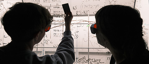 Two students examine chemistry calculations on an illuminated white board.