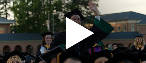 Several graduates stand and wave amongst the other seated graduates with a play icon overlayed to indicate video content