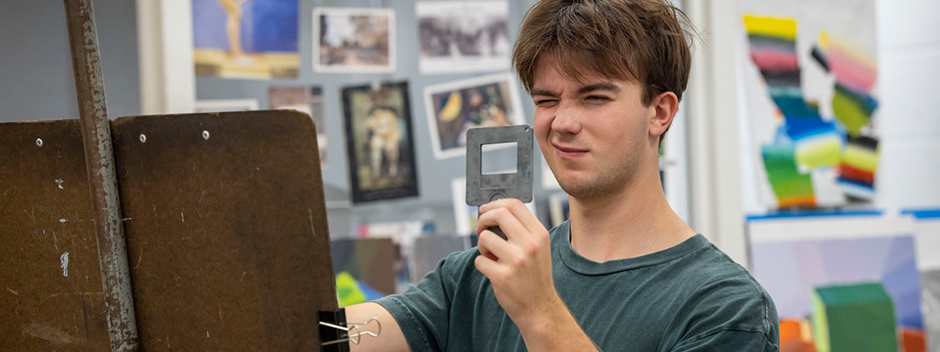 A student squints with one eye as they draw at an easel with many colorful drawings posted on the wall in the background
