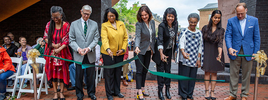 Several campus leaders smile as they cut a long green ribbon beneath the brick memorial