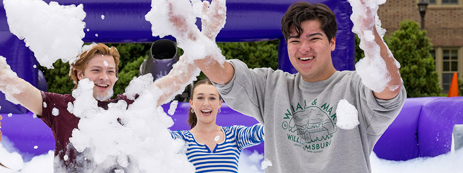 Three smiling students sweeping arms of bubbles up in the air within a large inflatable in the Sunken Garden at the carnival