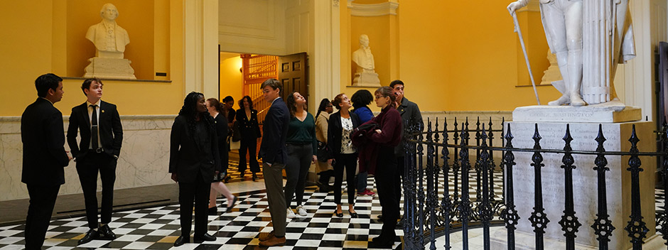 A group of students in professional attire gather in the yellow-walled rotunda of the Virginia State Capitol