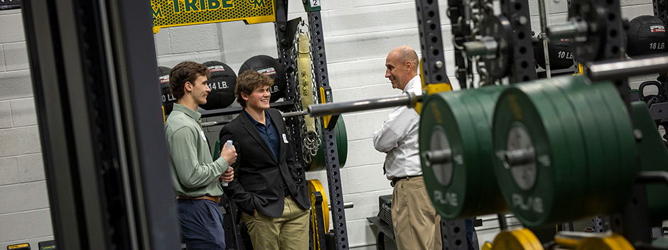 Three people smile and talk amongst strength and conditioning machines in the new Mackesy Sports Performance Center