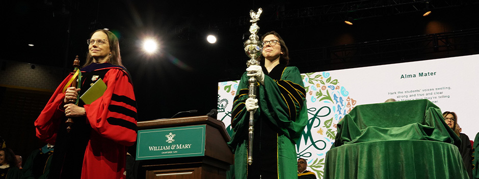 Ceremony marshals in full regalia stand at the front of the stage with the lyrics to the Alma Mater displayed on a large screen in the background 