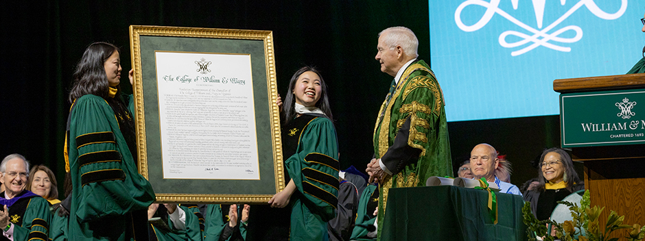People in green and gold robes hold up a large framed copy of William &amp; Mary's charter on stage in front of Chancellor Robert M. Gates.