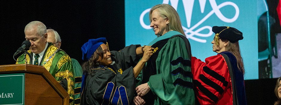 Chancellor Robert M. Gates and two faculty members attach a stole to the robes of Governor Abigail Spanberger on stage at the ceremony.