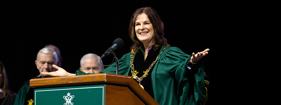President Katherine Rowe stands with open arms at a podium on stage at the ceremony with honored guests in the background.