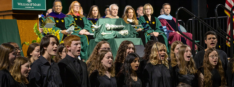 Students in black suits and dresses sing to attendees in front of the stage at the ceremony with honored guests and the school maces in the background.