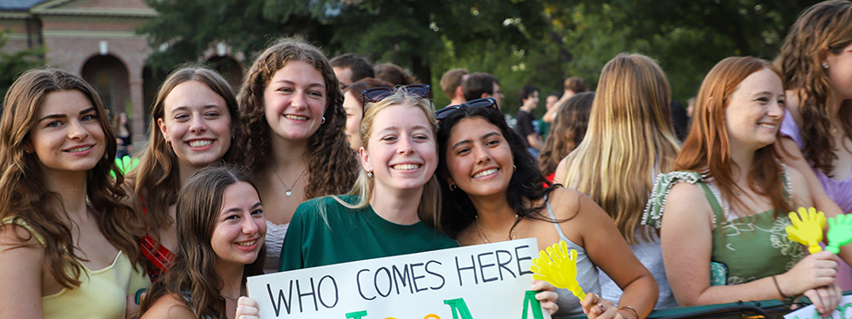 A group of smiling students gather together in the Wren Courtyard holding a sign that reads "Who comes here..."