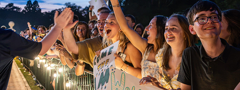 Students stand behind a barrier, smiling, waving signs and giving out high fives to passing students at an outdoor evening event with bright lights in the foreground.