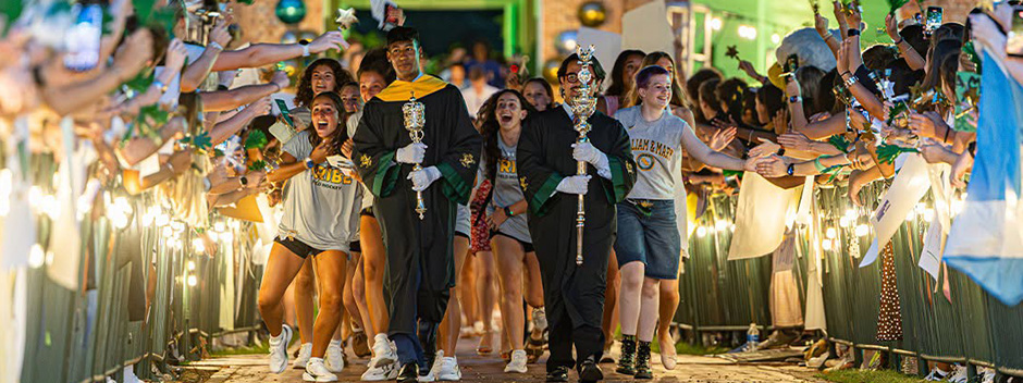 Student leaders in regalia, holding large symbolic maces, lead a cheering column of students through cheering members of the community