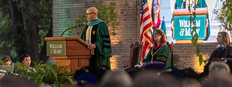 Convocation speaker Michael K. Powell, President Katherine A. Rowe and Provost Peggy Agouris on an outdoor platform in the Wren Yard at night while Powell speaks from a podium.