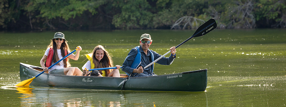 Three smiling people wearing life vests and W&amp;M hats are rowing a canoe on Lake Matoaka
