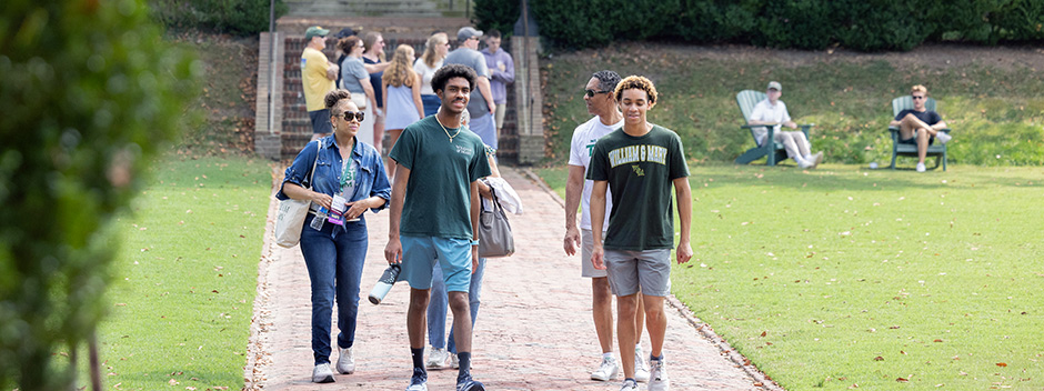 A family in W&amp;M attire walks together across the Sunken Garden with other families gathered in the background