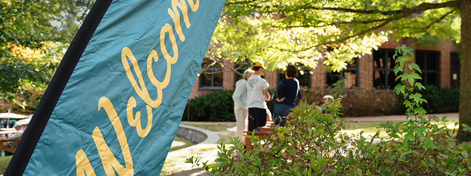 A welcome sign in the foreground says Welcome and a small group of people are in the background surrounded by sunshine and tree foliage beginning to show signs of fall colors.