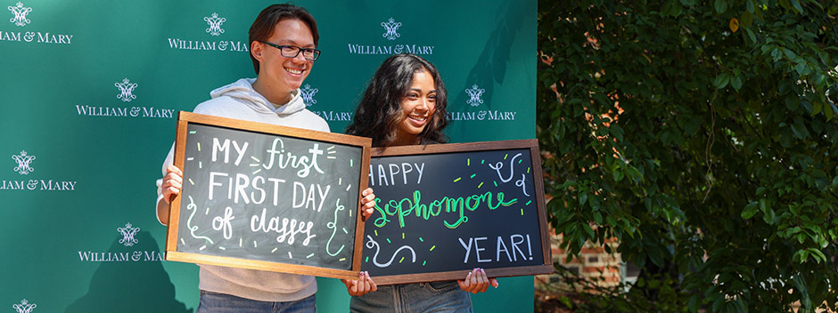 Two students posed smiling in front of a sign with many William &amp; Mary logos holding chalkboard sign that read "My first First Day of Classes" and "Happy Sophomore year" 