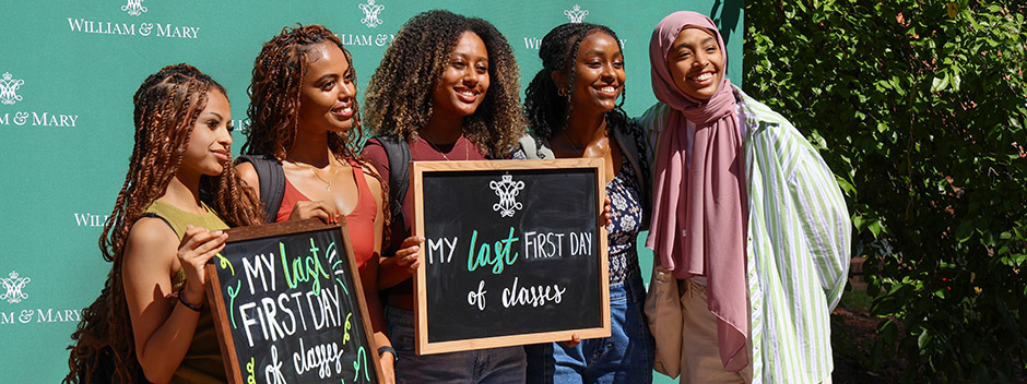Five students posed smiling in front of a sign with many William &amp; Mary logos holding chalkboard signs that read "My last First Day of Classes"