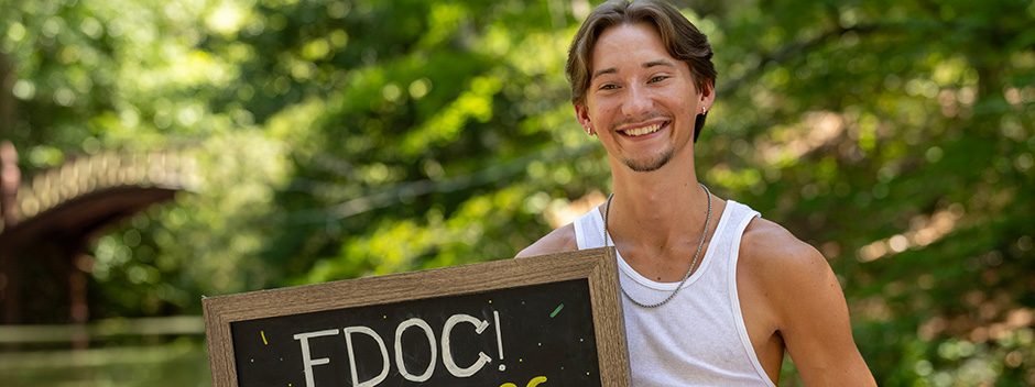 A smiling student holding a chalkboard sign with the lettering FDOC, with the Crim Dell and bridge in the background