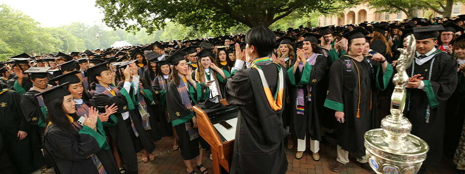 Large crowd of students in graduation attire gather to hear a student speaker at a podium in the Wren Yard