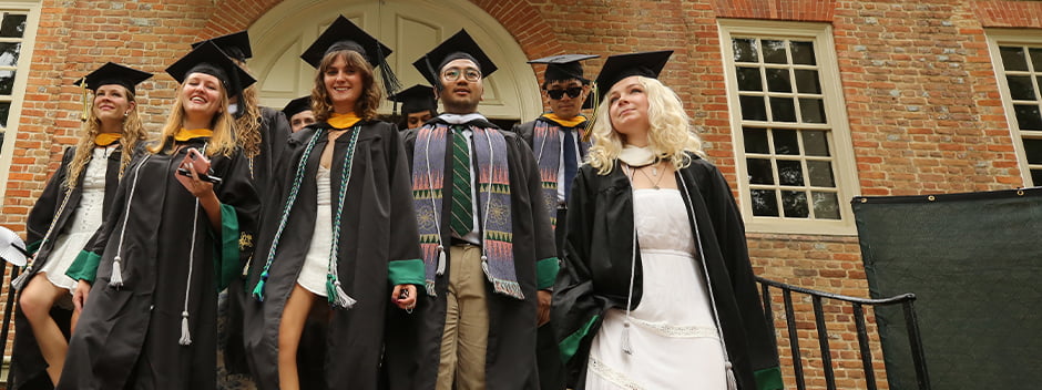 A group of smiling graduates in commencement attire walk out of the Wren Building