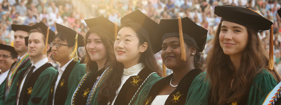 A row of smiling graduate students in full regalia with families and guests filling the stadium seating in the background