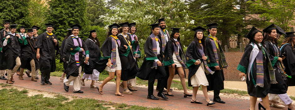 Students in Commencement attire smiling and walking along the Sunken Garden on the brick walkway towards the Commencement ceremony
