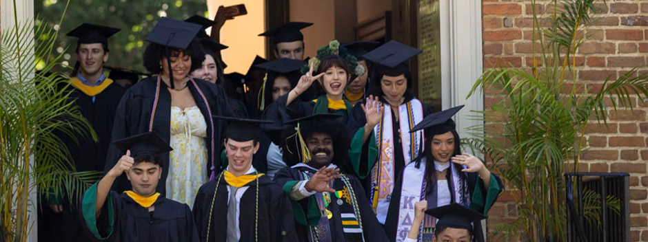 A line of graduates wave and smile as they exit an historic brick building 