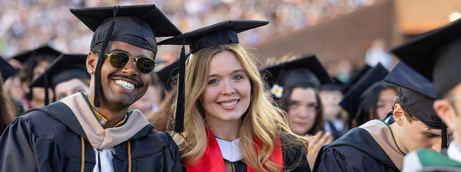 Two smiling graduates in full regalia pose for the camera while seated at the main ceremony on the field of Zable Stadium