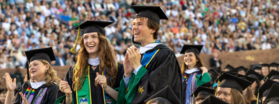 Smiling graduates stand amongst other graduates on the field of Zable Stadium during the main ceremony