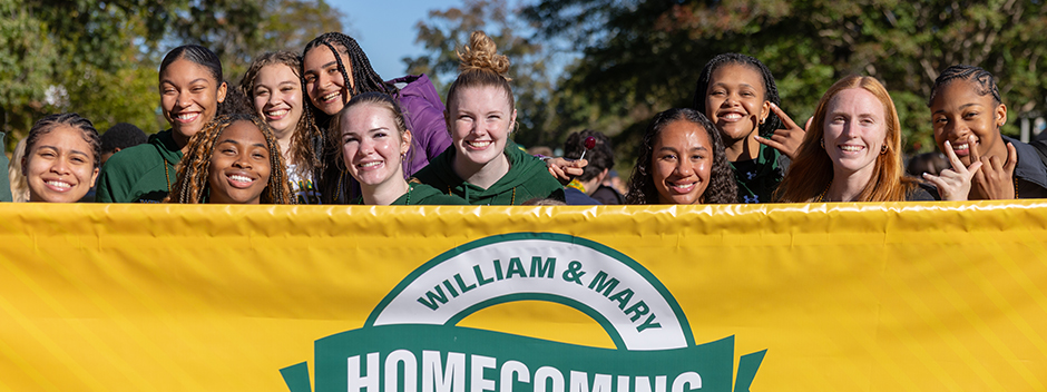 Smiling students walk behind a large yellow Homecoming banner at the parade