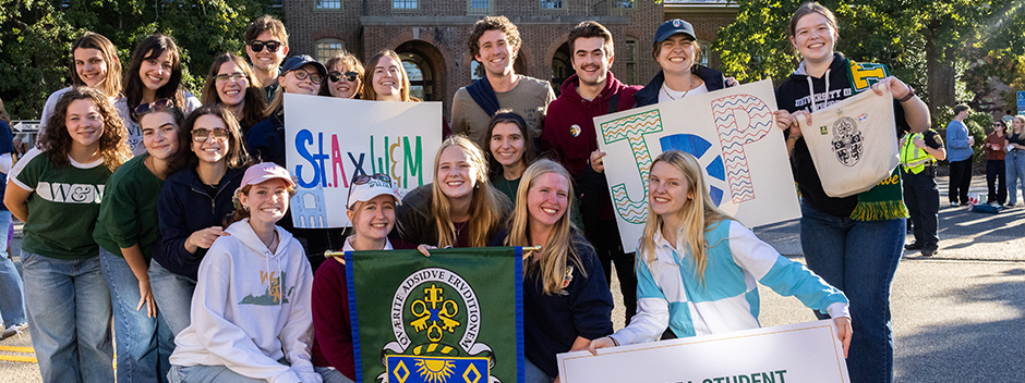 A group of smiling students pose with St Andrews Joint Degree Programme signs on campus