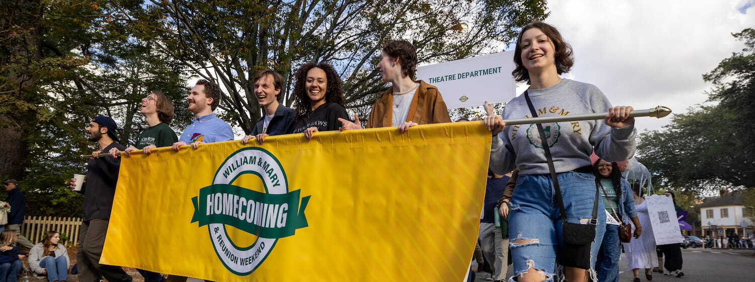 A bright yellow Homecoming &amp; Reunion Weekend banner is carried by a row of smiling people in the parade with a Theatre Department sign and more parade participants in the background