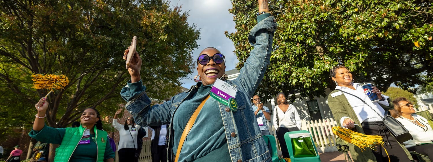 A smiling W&amp;M alumnus in sunglasses and a denim jacket waves both arms in the air while holding a phone.