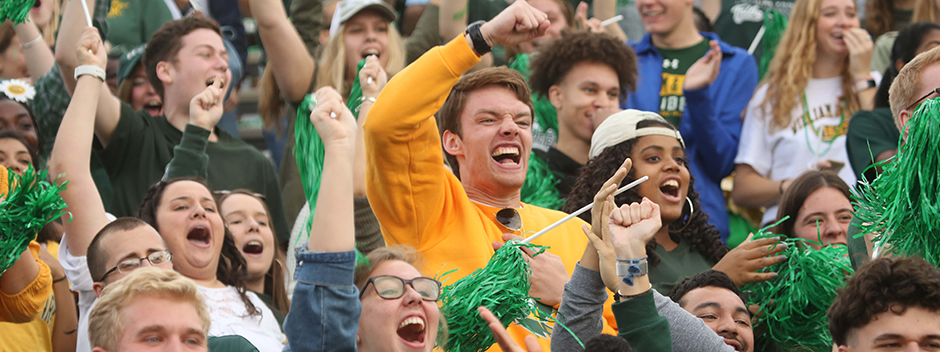 A crowd of enthusiastically cheering students in green and gold attire with green pompoms in the stands of Zable Stadium