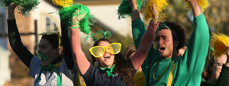 Several students wearing green and gold clothes, giant sunglasses and pompoms cheer on top of a float on Richmond Road