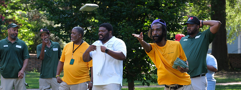 A group of employees gathered watching someone toss a beanbag in a cornhole tournament