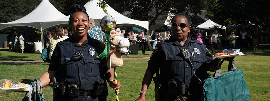 Two smiling W&amp;M Police officers holding holding birthday decor and food at a celebration in the Wren Yard