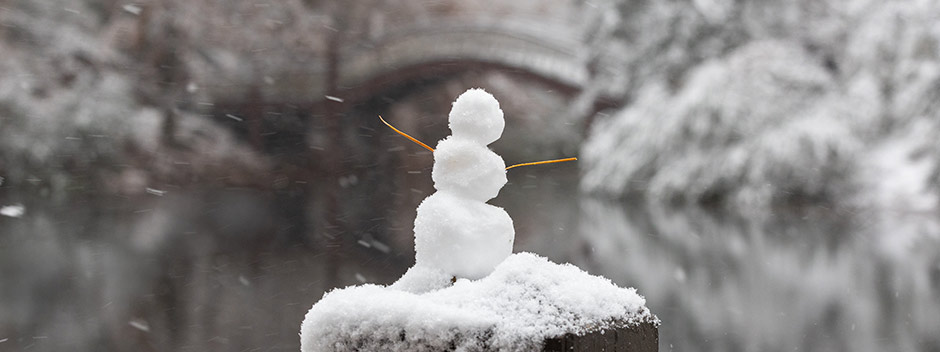 A small snowman with twigs for arms atop a snow-capped post with the Crim Dell bridge in the background