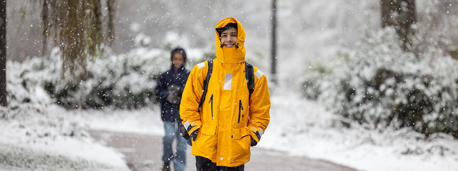 A smiling student bundled in a bright yellow winter jacket walks along a brick path as it snows