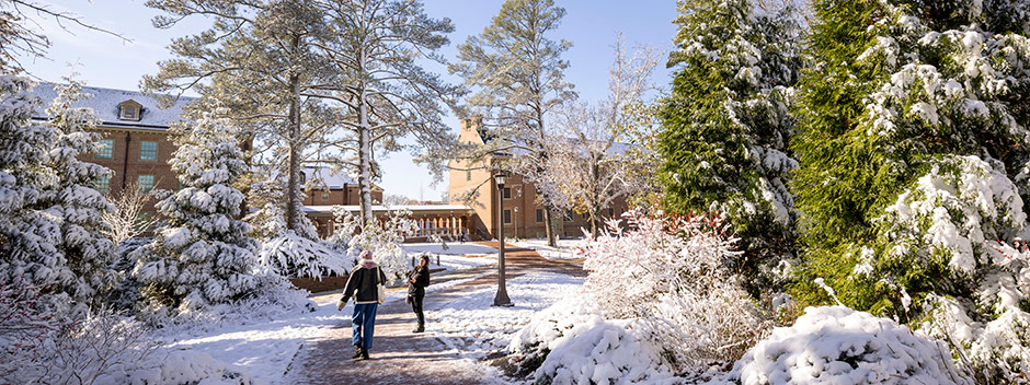 Two students bundled in winter attire walk along a curvy brick path with snow-blanketed trees and buildings around them