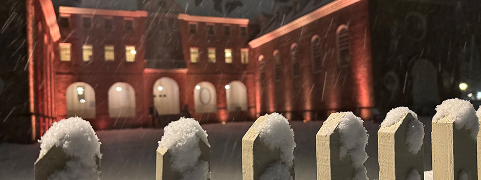 Snow accumulating along the white pickets of the Wren Courtyard in the foreground with a warmly lit Wren Building in the background