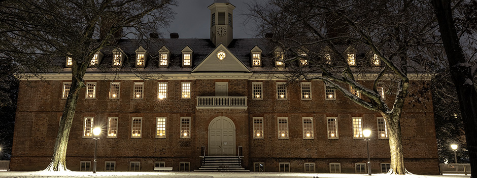 A front view of the historic Wren Building at night with a light blanket of snow on the building, grass and trees, and a candle lit in each window for the holidays