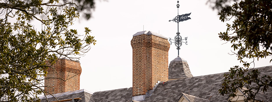 Top of the historic Wren Building including two brick chimneys and the 1693 weather vane viewed through evergreen tree foliage on a gray-sky day