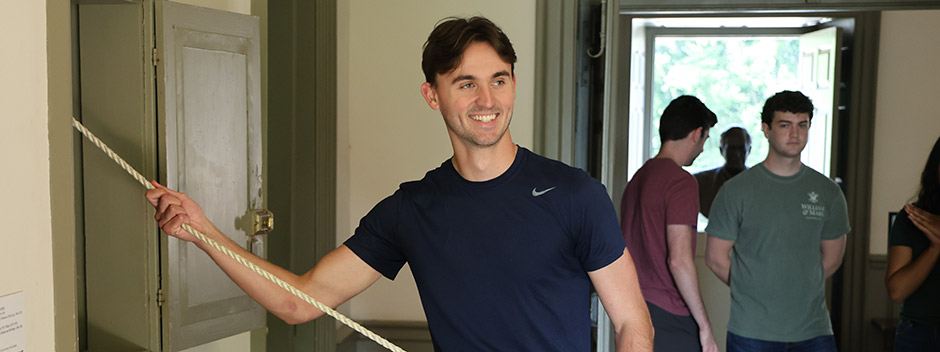A smiling student poses while pulling a rope in the Wren Building to ring the Wren Bell