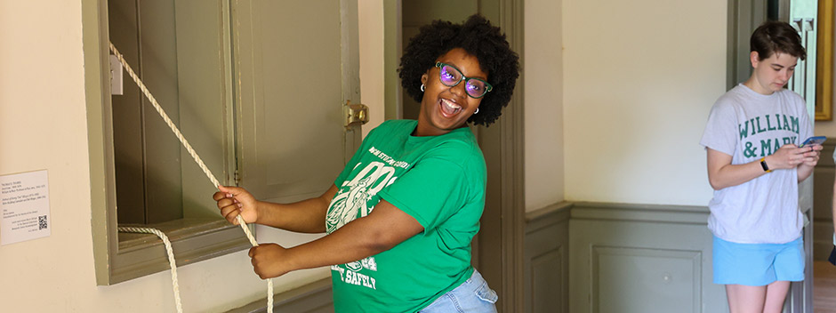 A smiling student poses while pulling a rope in the Wren Building to ring the Wren Bell