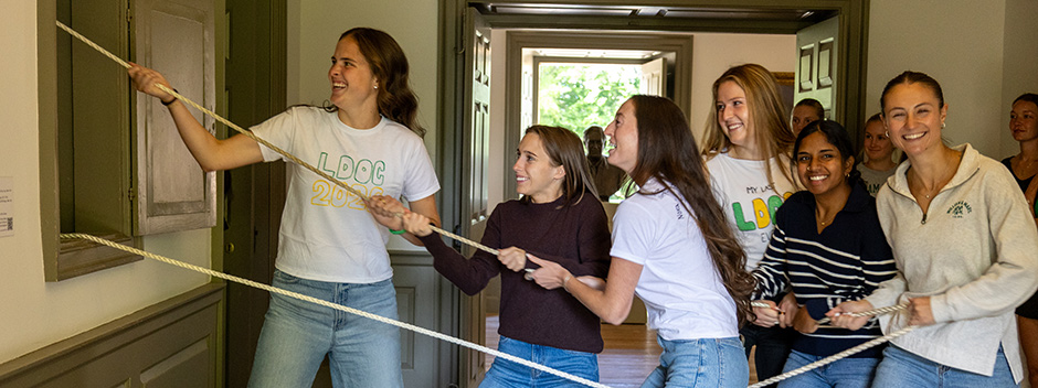 Six smiling students pull a rope together in the Wren Building to ring the Wren Bell