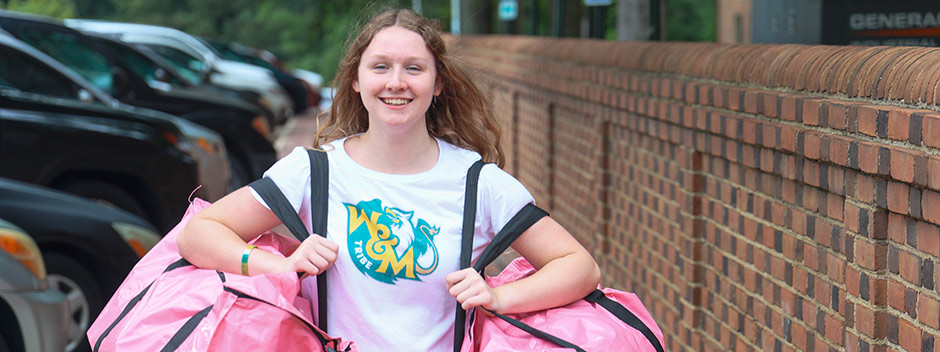 A smiling student wearing a W&amp;M Tribe T-shirt carries two large pink bags along brick walkways and rows of cars