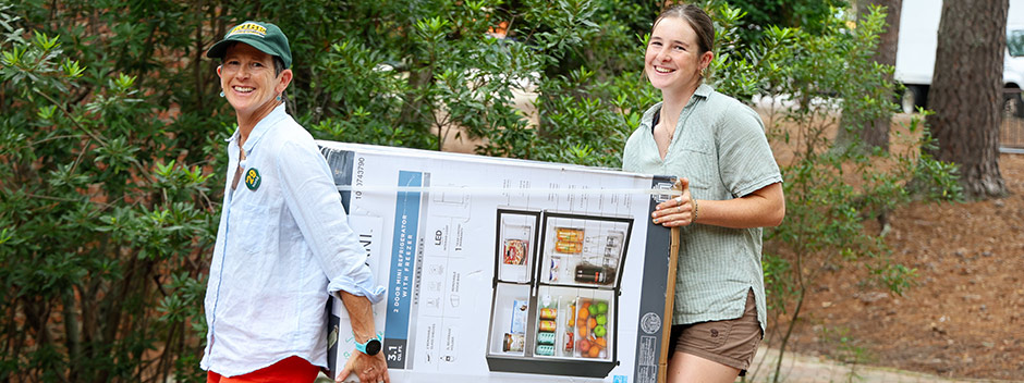 A smiling family member and new student carry a boxed refrigerator on campus