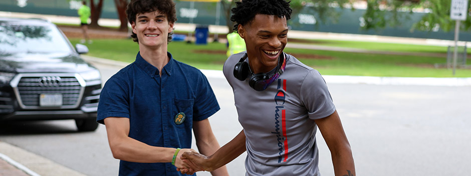 Two students shake hands and laugh on a campus walkway