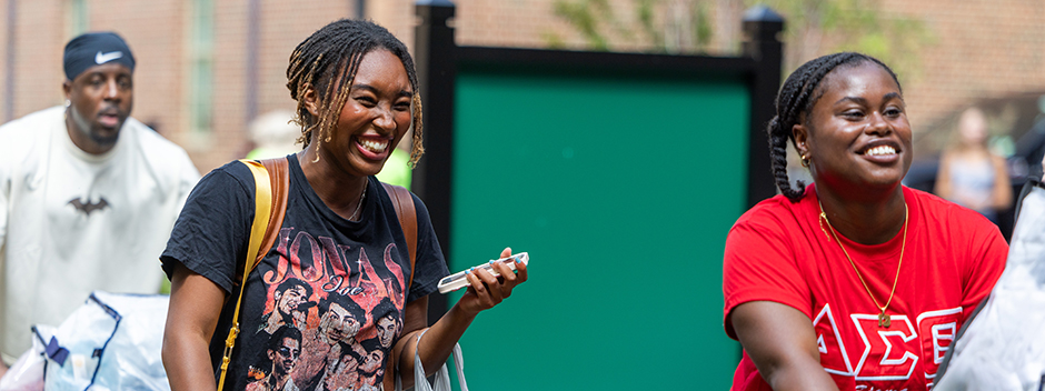 Two people smiling and carrying loads of belongings across campus